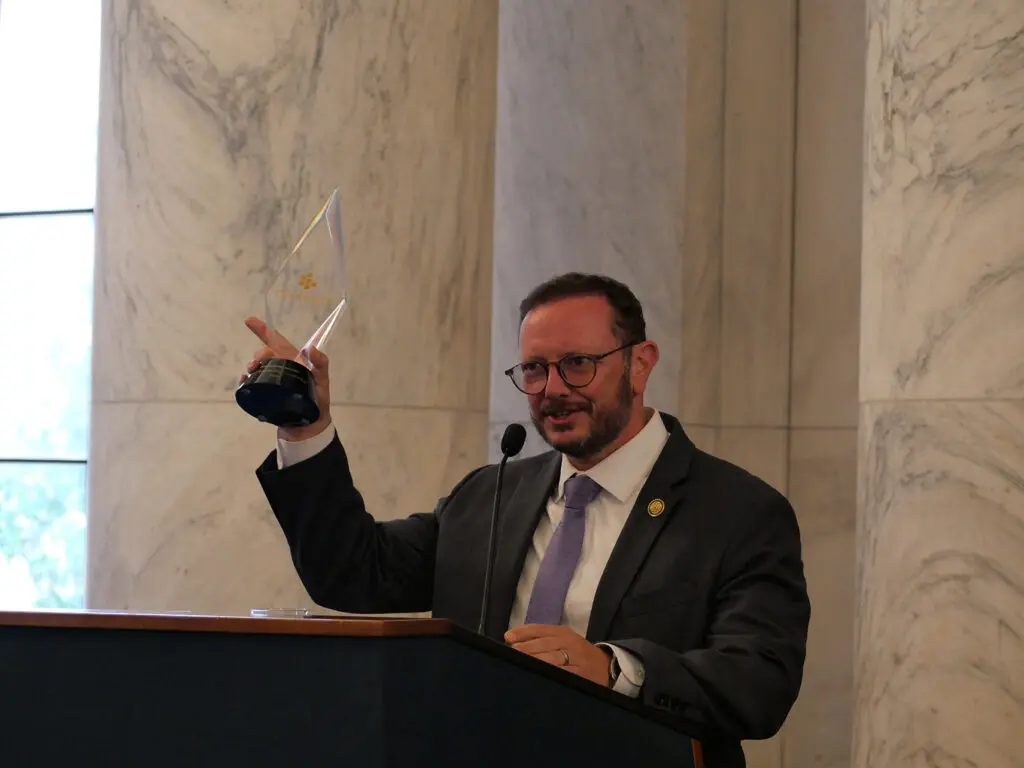 Man in a suit and glasses raises a glass award at a podium during a formal ceremony in a marble-walled room.