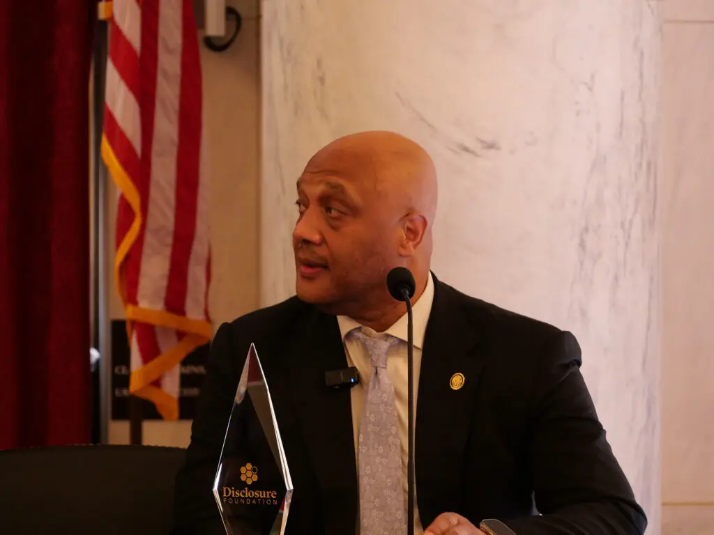 Man in a dark suit and light patterned tie speaks at a podium with a microphone; a glass award labeled 'Disclosure Foundation' stands in front, with an American flag in the background.