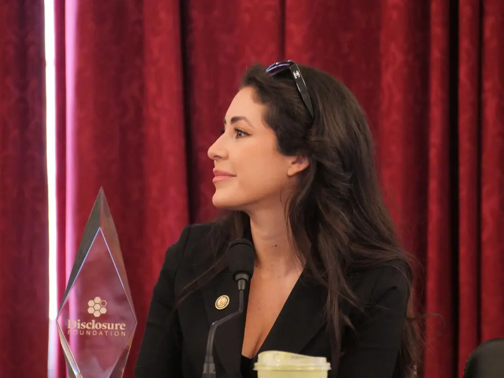 Side profile of a woman in a black blazer speaking at a panel, with a glass award that reads 'Disclosure Foundation' and red velvet curtains behind her