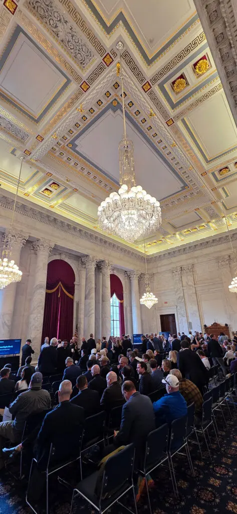 Grand ornate hall with a large crystal chandelier, marble columns, and red-draped windows; attendees seated in rows for a formal event.