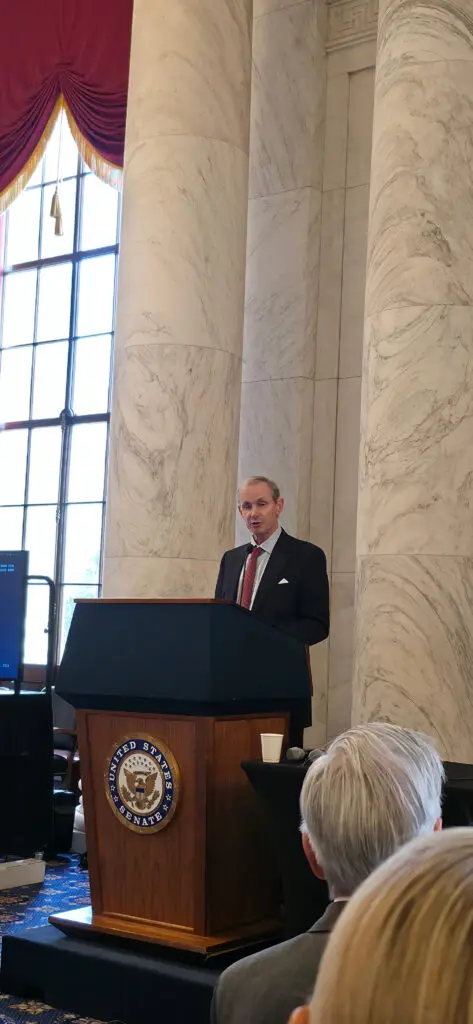 Man in a dark suit and red tie speaks at a wooden podium with the U.S. Senate seal, marble columns in the background.
