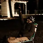 Man in a dark jacket sits in a folding camping chair at night, raising his right hand with one finger pointed up.