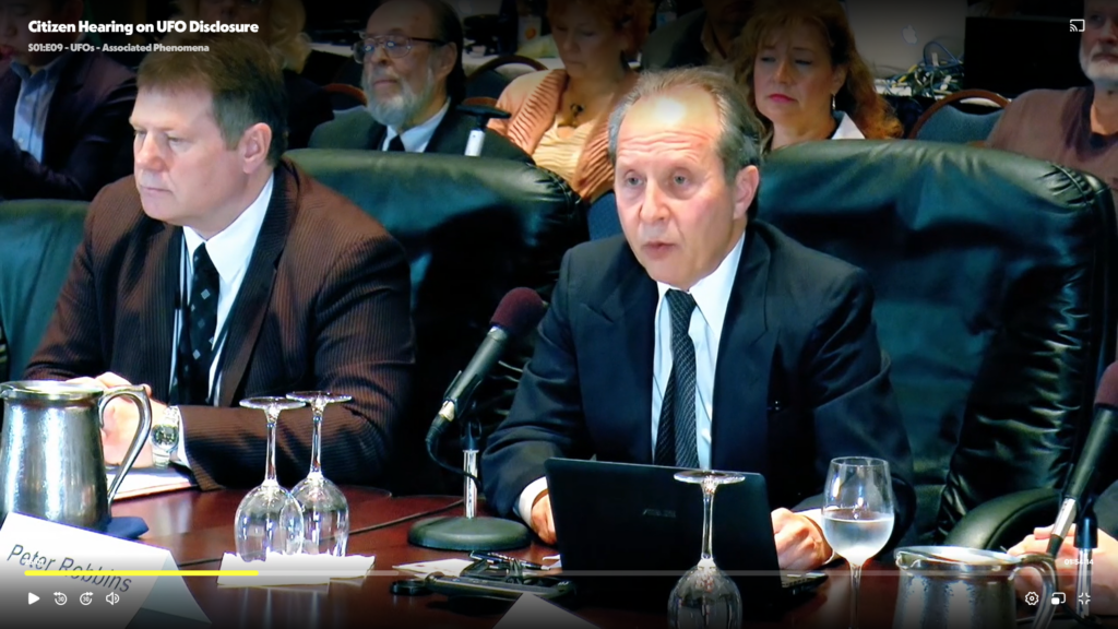 Two suited panelists sit at a conference table with microphones and glasses, addressing an audience in the background.