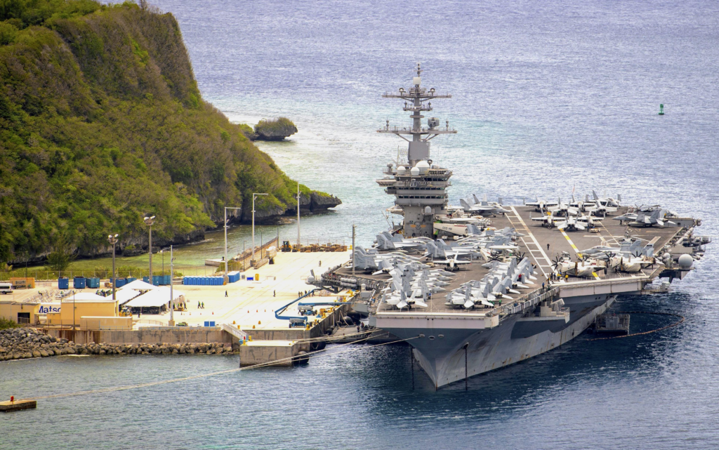 image Aircraft carrier docked at a harbor with numerous fighter jets on its deck and a rocky, forested coastline nearby.