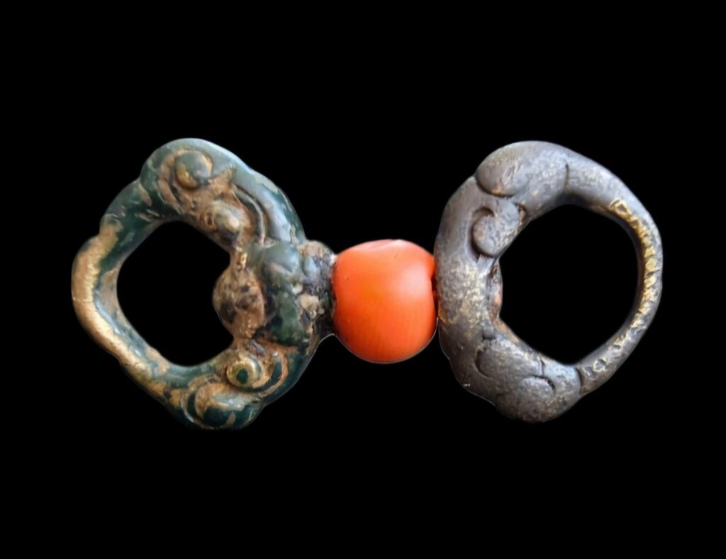 1-thogcha-amulet - UAPedia Close-up of two aged, irregular metal rings connected by a bright orange bead in the center against a black background.