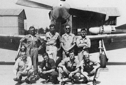 Group of eight men in flight suits posing in front of a propeller-driven bomber on a tarmac at an airbase. (informational)
