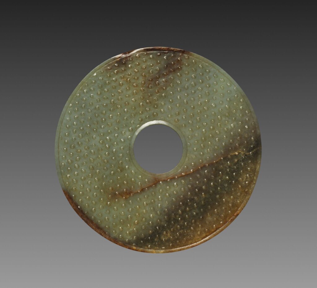 Jade Bi Disc Dropa disc Heritage - UAPedia Close-up of a greenish circular metal plate with a central hole and many raised rivets; rust along the bottom edge against a dark gradient background.