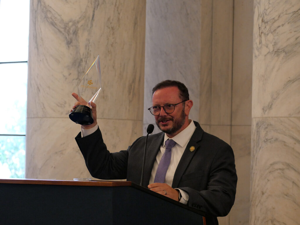 Man in a suit and glasses raises a glass award at a podium during a formal ceremony in a marble-walled room.
