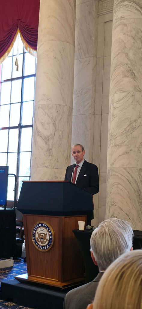 Man in a dark suit and red tie speaks at a wooden podium with the U.S. Senate seal, marble columns in the background.