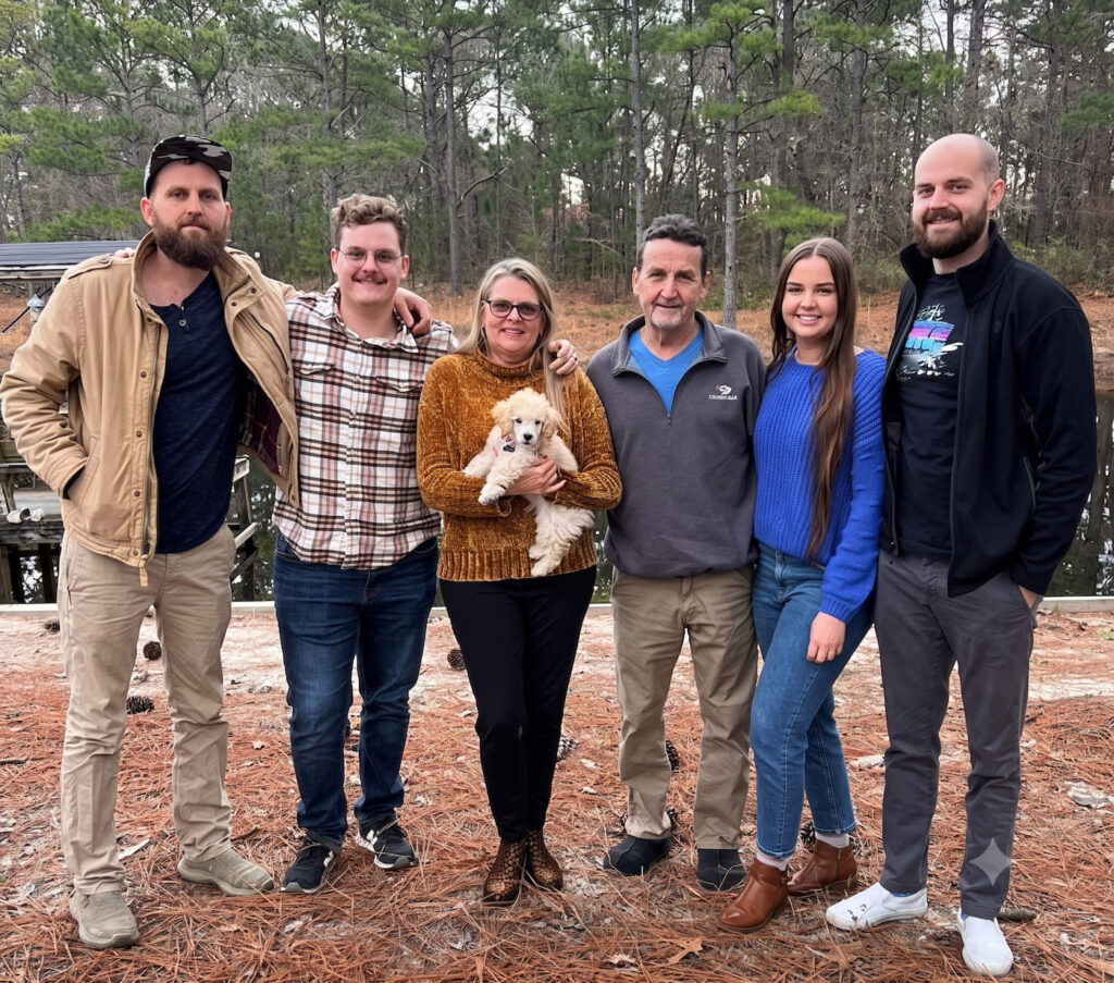 Six adults stand arm-in-arm outdoors on a pine-needled ground, a woman in a mustard sweater holds a fluffy white puppy in front of them.