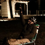 Man in a dark jacket sits in a folding camping chair at night, raising his right hand with one finger pointed up.