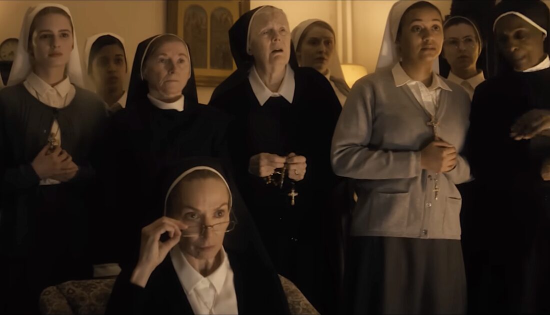 Screenshot An elderly nun sits in the foreground, adjusting her glasses while a group of nuns in habit stand behind her, some holding rosaries.