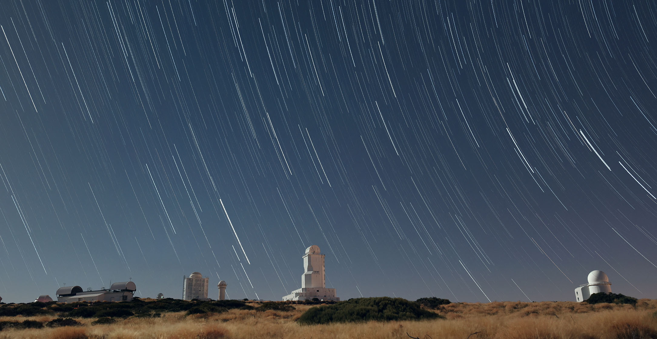 evanto-spain-tenerife-star-trails-over-teide-observator-2026-03-09-06-02-59-utc-33p Long-exposure night scene of star trails over a row of observatories on a hill.